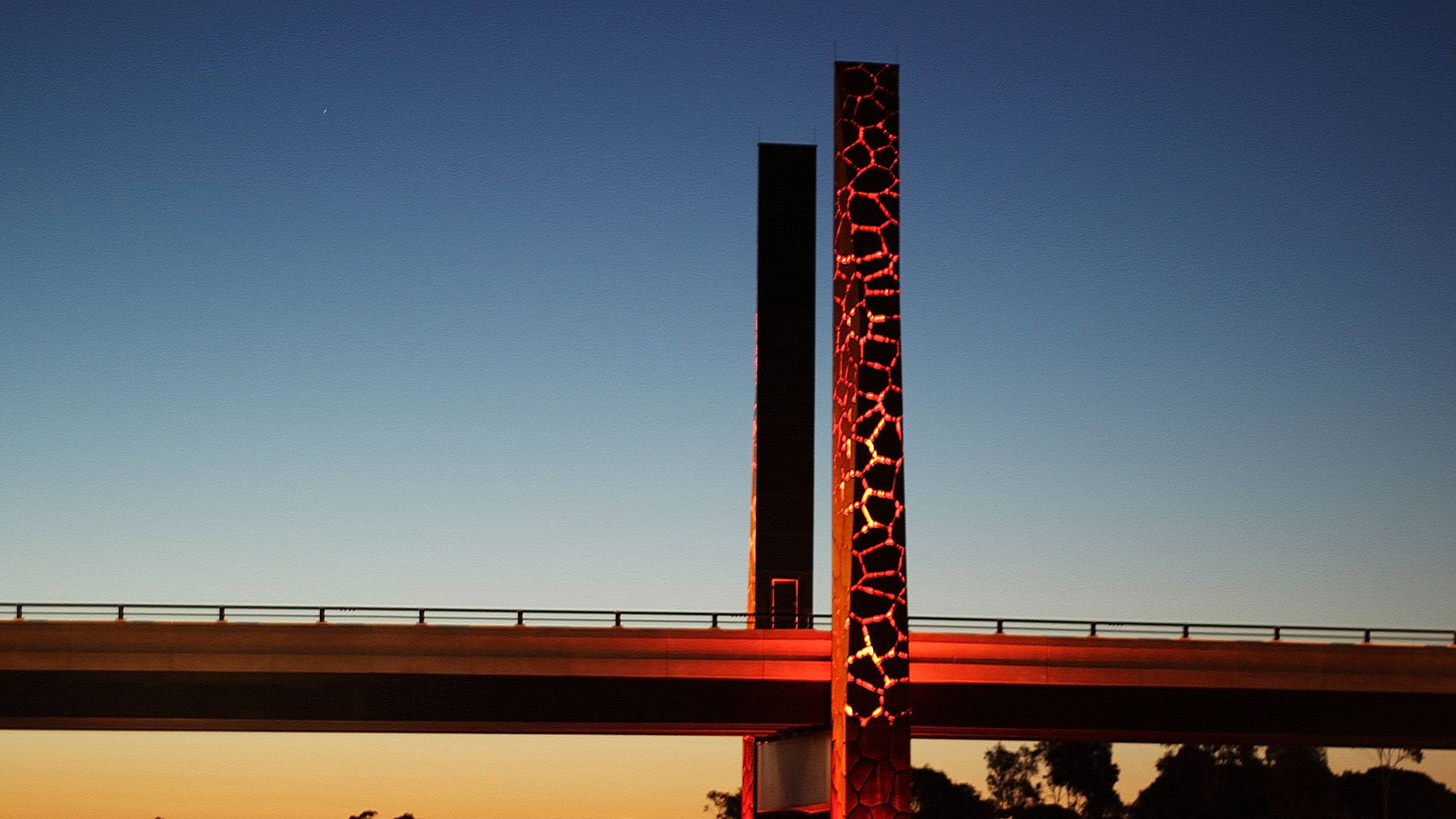 An installation of high-power linear luminaires inside the two mast features of the bridge. The luminaires create a glowing effect from inside the structure.

